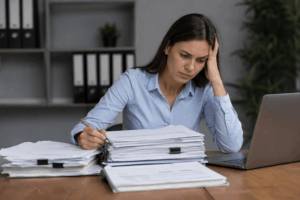 Woman in a blue shirt leans on her head while reviewing a large pile of papers at a desk with a laptop nearby.